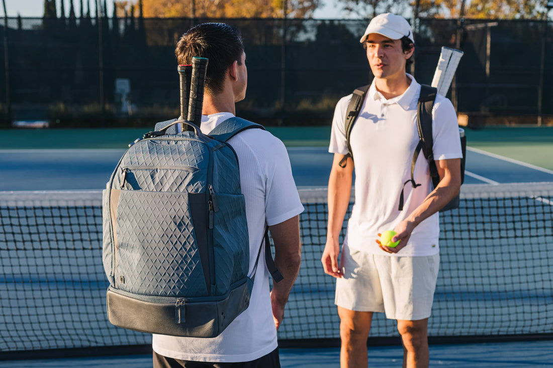 Two men talking to each other on a tennis court. One man is carrying the NYC backpack from Doubletake.