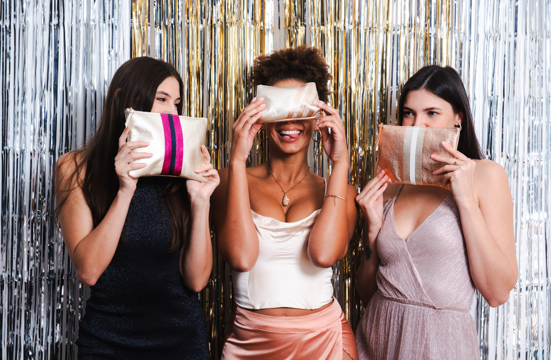 Three women dressed in elegant outfits hold small metallic clutches over their faces, laughing. The background is decorated with silver and gold tinsel streamers.